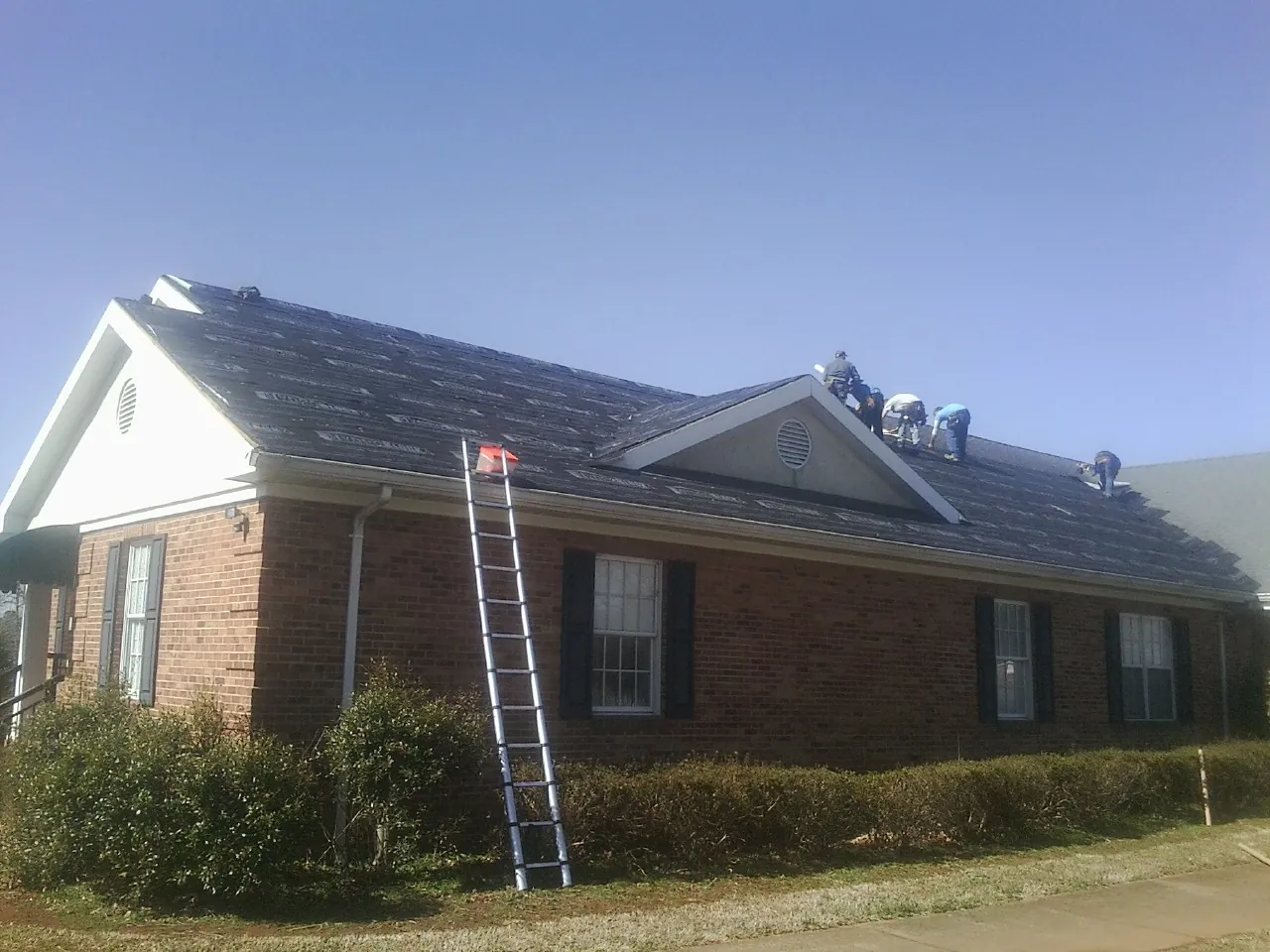 Crew tearing shingles off a brick ranch roof with a ladder set — Metal Roof Repair work by our crew in Deanville