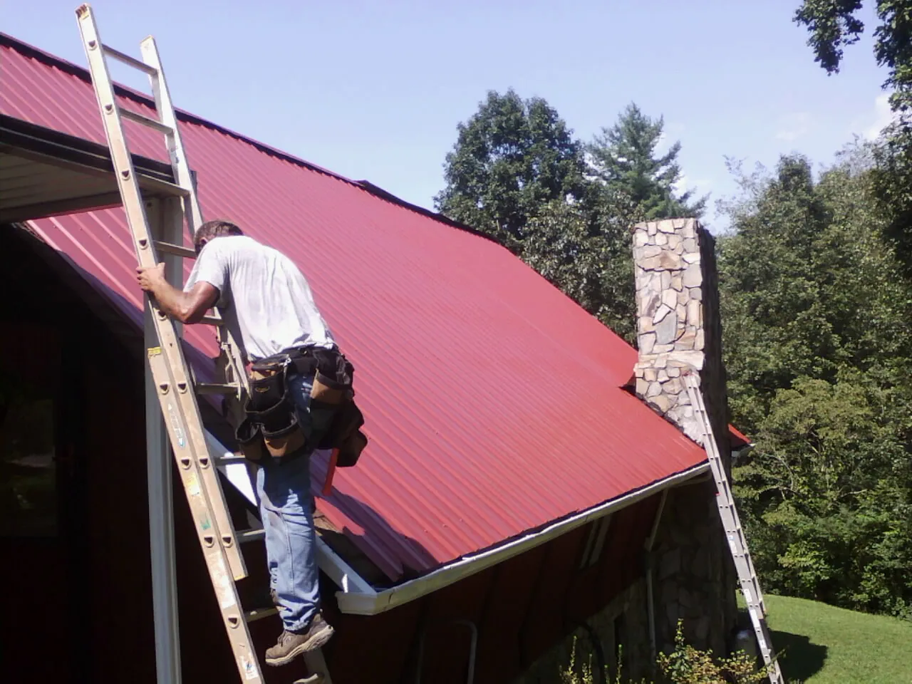 Roofer on a ladder beside a red metal roof and stone chimney — Soffit & Fascia Repair by Roofing Brothers in Deanville